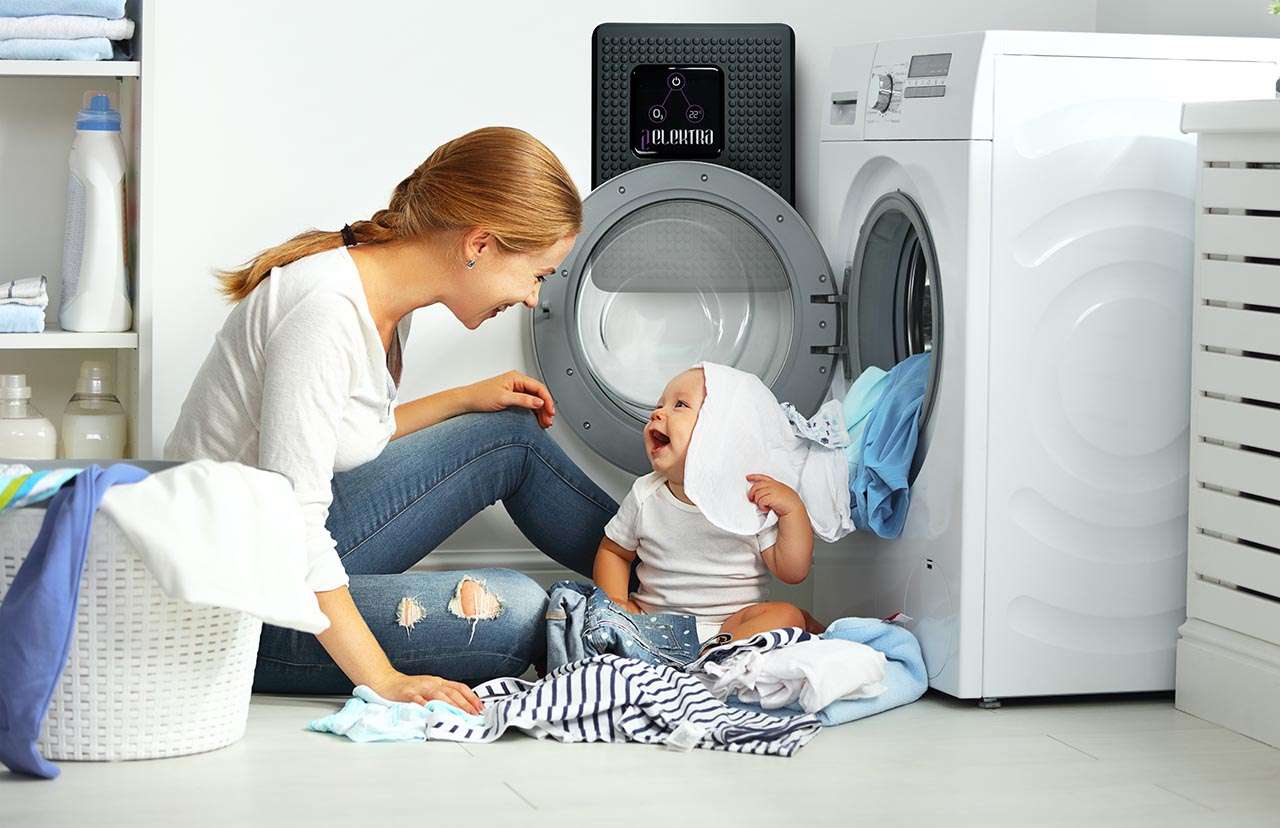 Mãe e bebé brincam junto a uma máquina de lavar aberta numa sala iluminada, rodeados de roupa limpa. A mãe sorri enquanto o bebé carrega uma peça de roupa na cabeça, criando uma atmosfera de felicidade e cumplicidade no lar. Atrás da porta da máquina de lavar roupa vê-se um aparelho ELEKTRA by ECOFROG.