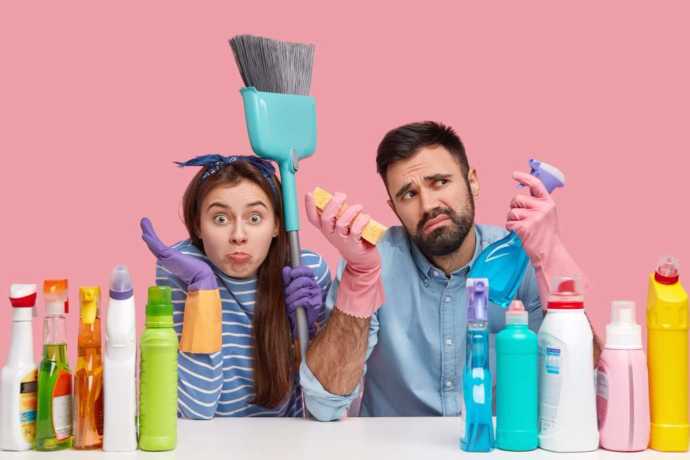 Um casal com luvas de plástico nas mãos, rodeado de produtos químicos e utensílios de limpeza, com um ar duvidoso e desanimado.