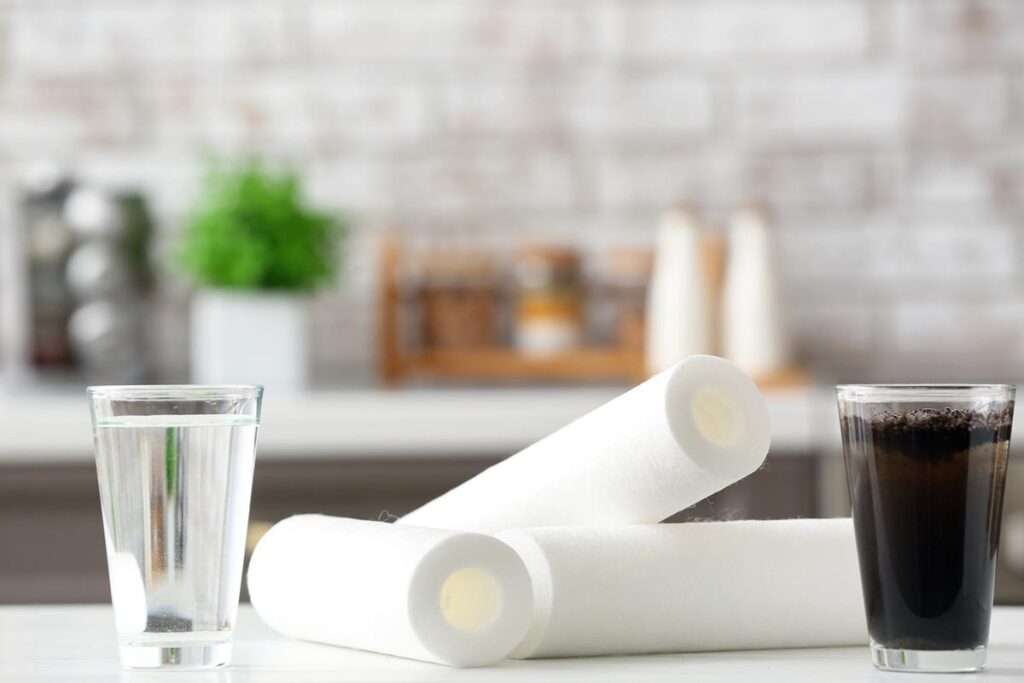 Numa cozinha, sobre uma bancada branca, há dois copos, um com água limpa e outro com água suja, com filtros de osmose no meio.