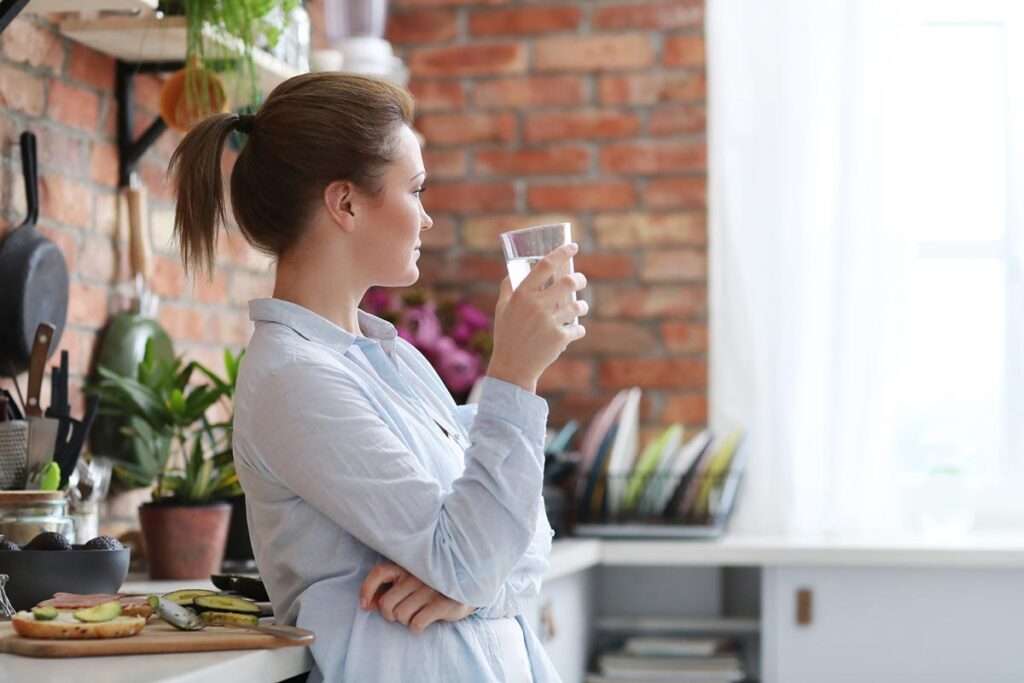 Mulher de aspeto descontraído numa cozinha a beber água filtrada por osmose e com alimentos saudáveis numa bancada ao seu lado.