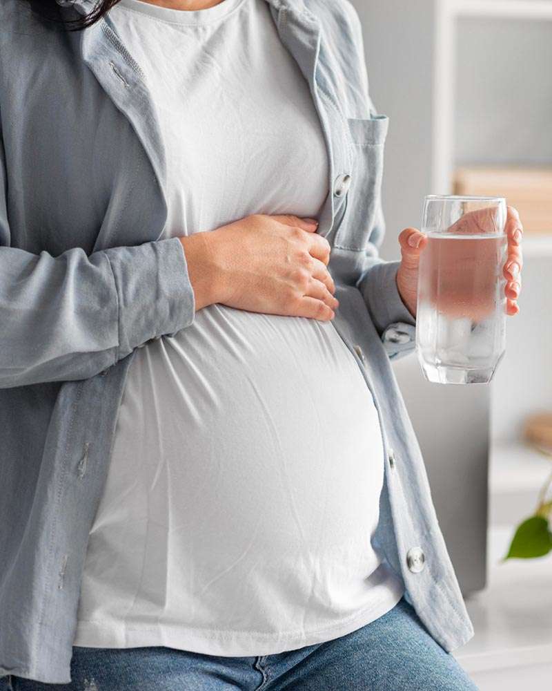 Mulher grávida a tocar na barriga, segurando um longo copo de água. Imagem muito brilhante com tons claros.