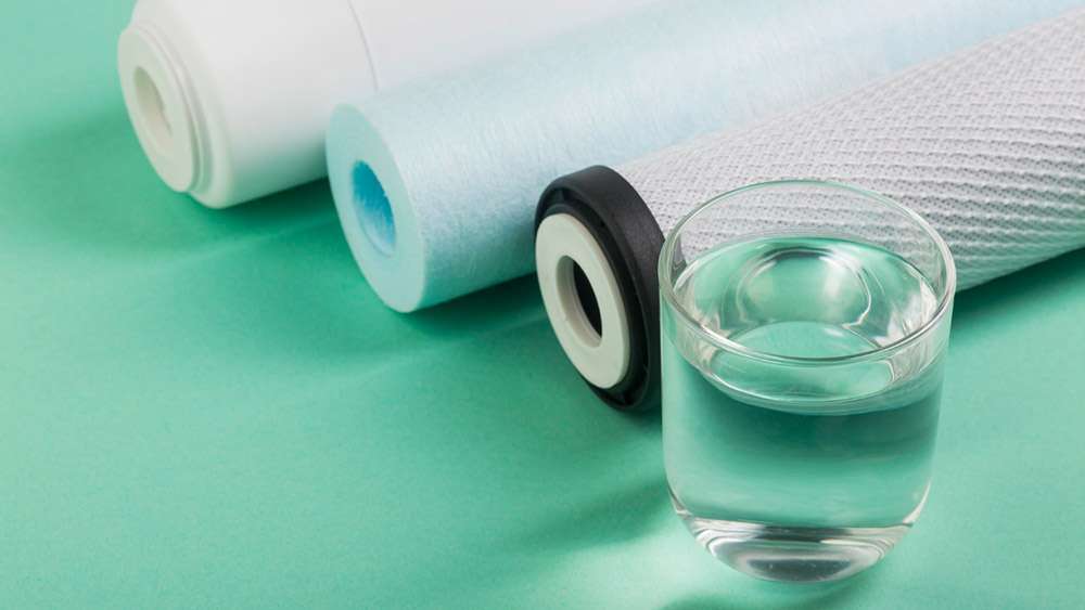 Três tubos de filtro de osmose diferentes ao lado de um copo cheio de água que repousa numa superfície verde menta.