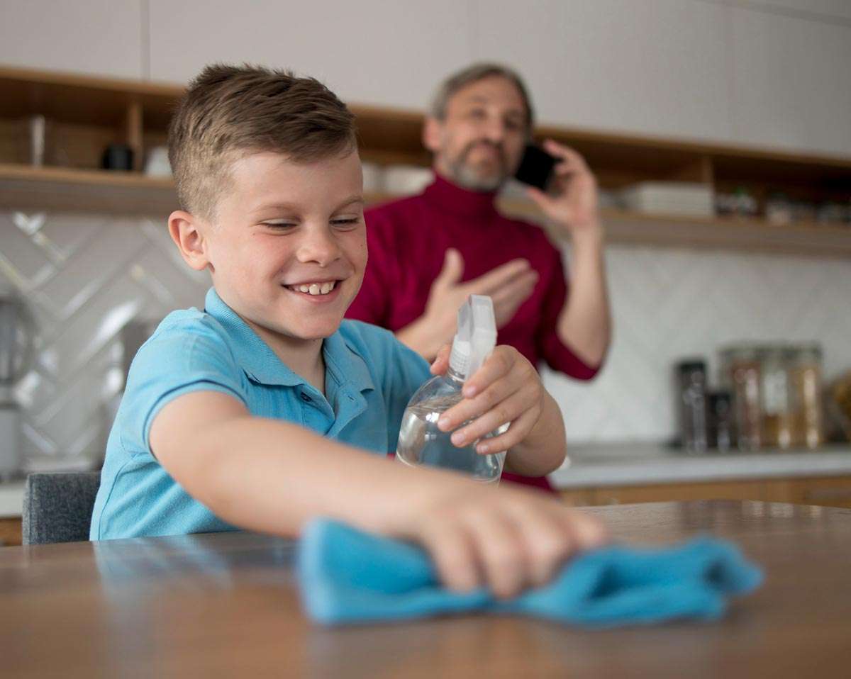 Numa cozinha, um pai fala ao telemóvel enquanto o filho limpa uma mesa com uma esfregona e um spray com água ozonizada.