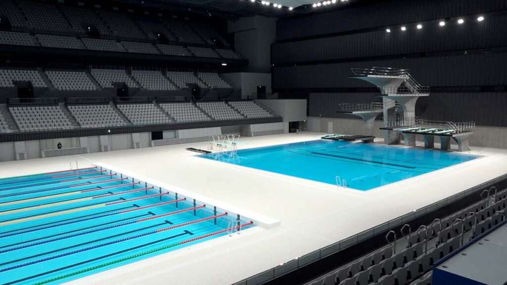 Vista panorâmica de uma piscina de competição desportiva e das pranchas de mergulho. Ao fundo, vêem-se as bancadas para o público.